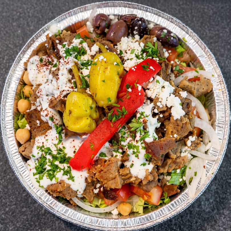 A close-up of a gyro bowl with meat, lettuce, tomatoes, chickpeas, peppers, feta, and sauce.