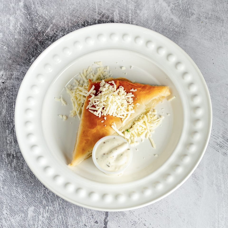 A golden-brown pastry triangle topped with shredded cheese and a side of creamy sauce on a white plate.