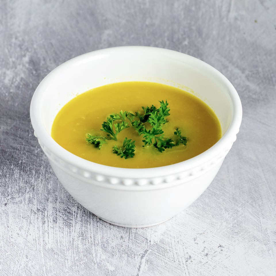 A white bowl filled with vibrant yellow soup, garnished with fresh green parsley, on a gray textured surface.