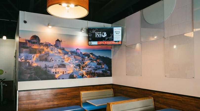 Restaurant interior with a large mural of a Greek village at sunset, booths, and a TV screen.