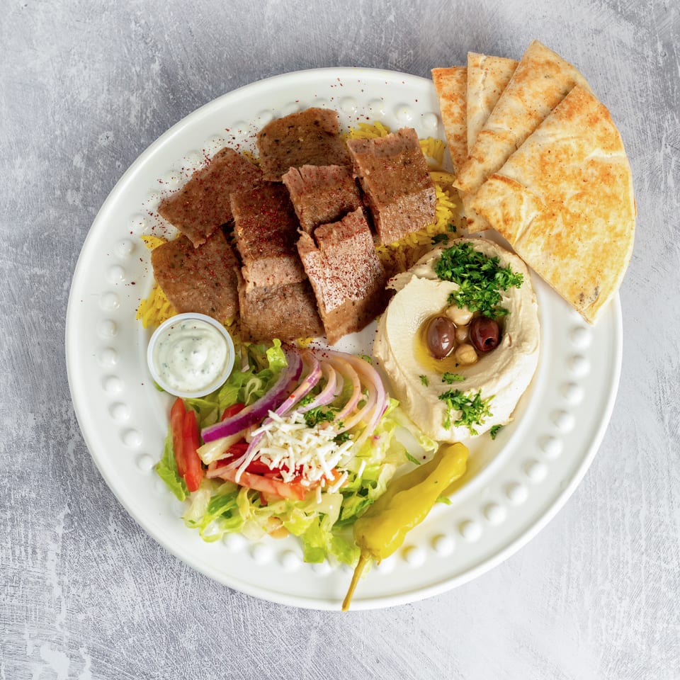 Gyro platter with sliced meat, yellow rice, hummus, pita bread, salad, and a side of tzatziki.
