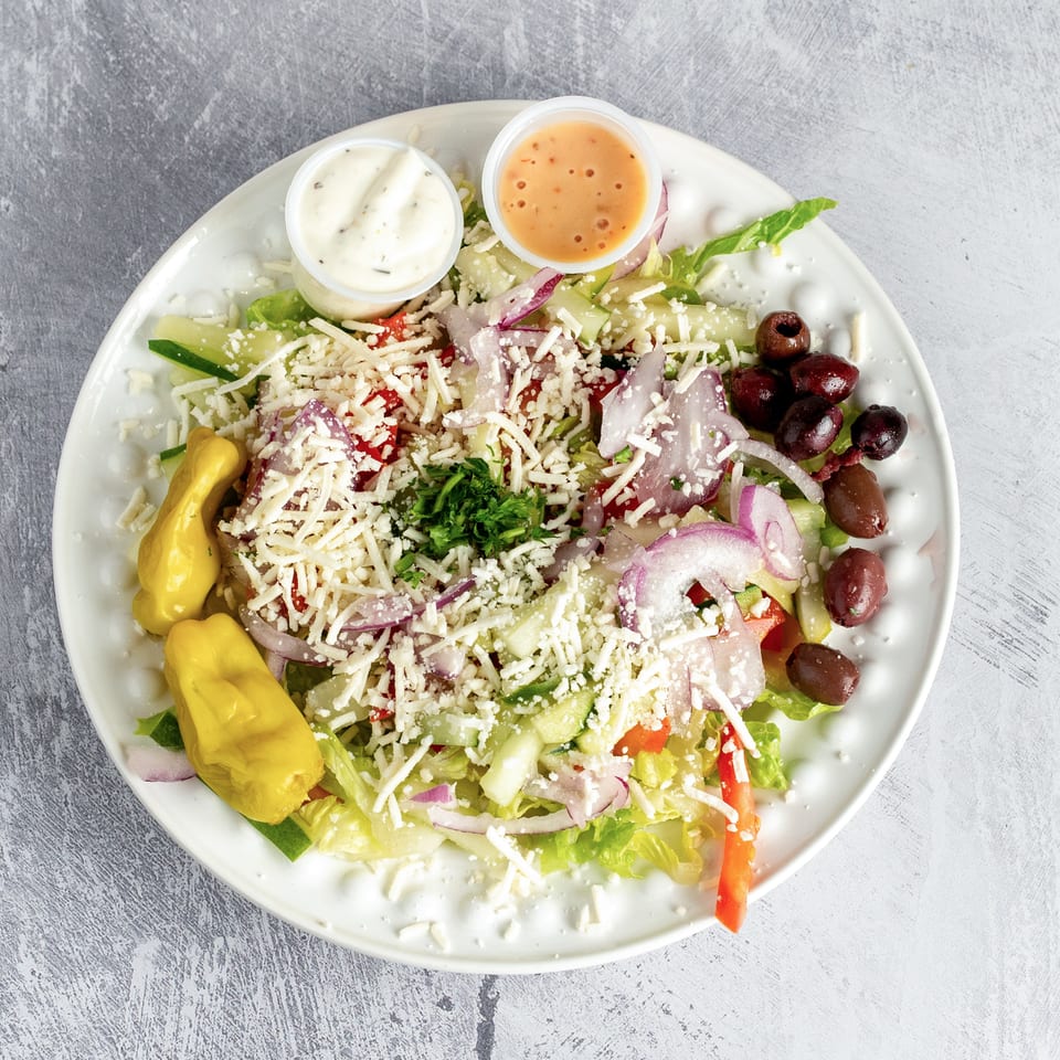Fresh garden salad with shredded cheese, red onions, olives, and two dressings on a white plate.