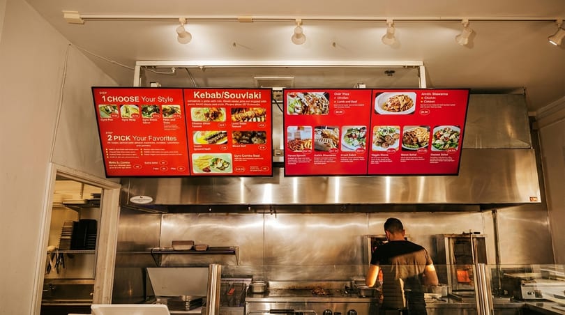 A chef cooks behind a counter with digital menu boards displaying various kebab and souvlaki dishes.
