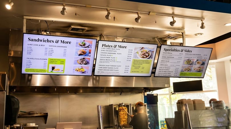 A restaurant counter with digital menu boards displaying Middle Eastern food options and a chef preparing shawarma.