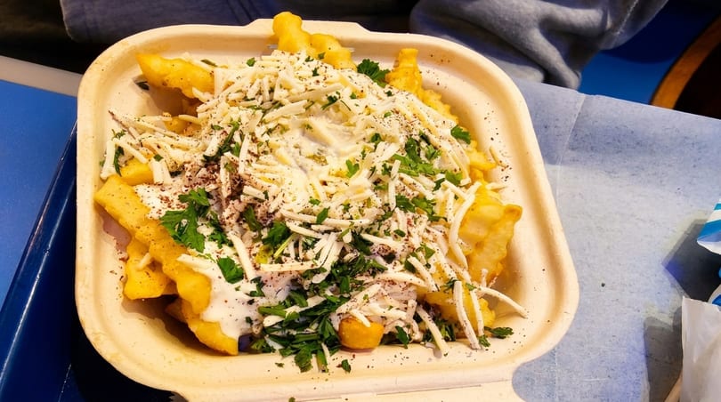Crinkle-cut fries topped with white sauce, shredded cheese, parsley, and spices in a takeout container.