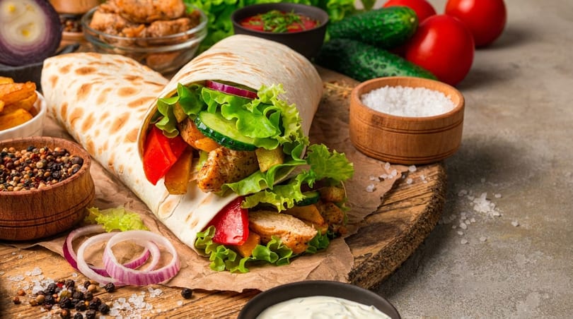 Chicken wrap with fresh lettuce, red peppers, and cucumbers, surrounded by spices and vegetables.