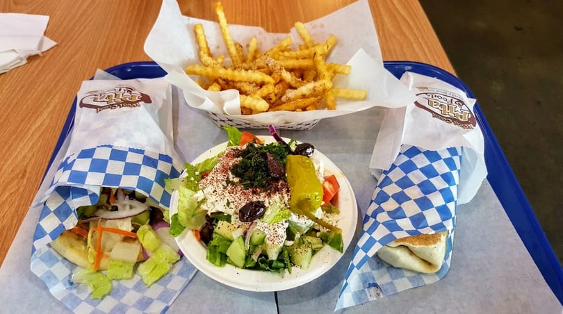 Two pita wraps, a side salad with feta and olives, and a basket of french fries on a tray.